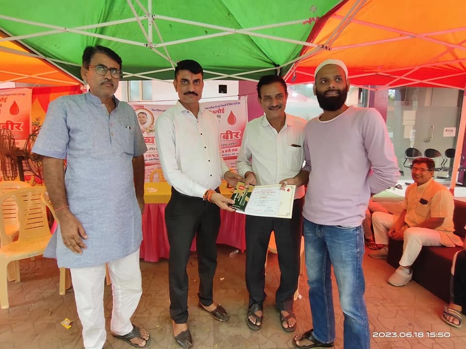 A donor is presented with a certificate under the green and orange canopies of the blood donation camp.