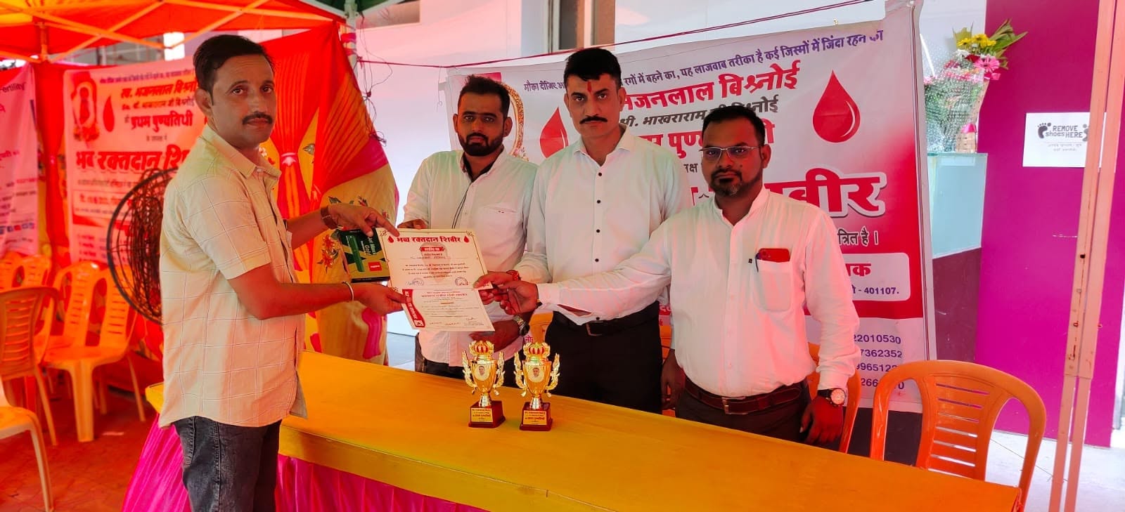 A certificate is handed to a donor across the registration table at the blood donation camp, with two trophies set out in front.
