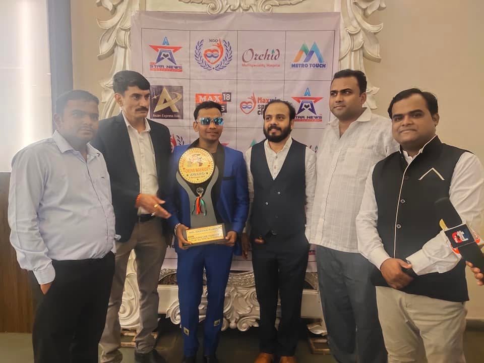 Six guests stand for a group photograph in front of a sponsor backdrop carrying the Orchid Multispeciality Hospital logo, one of them holding a Corona Warrior Award trophy.