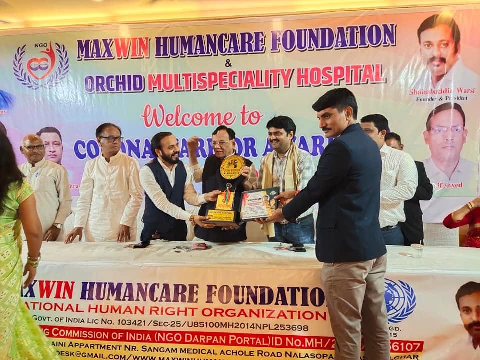 An award is handed across the head table beneath a banner reading "Maxwin Humancare Foundation & Orchid Multispeciality Hospital — Welcome to Corona Warrior Award".