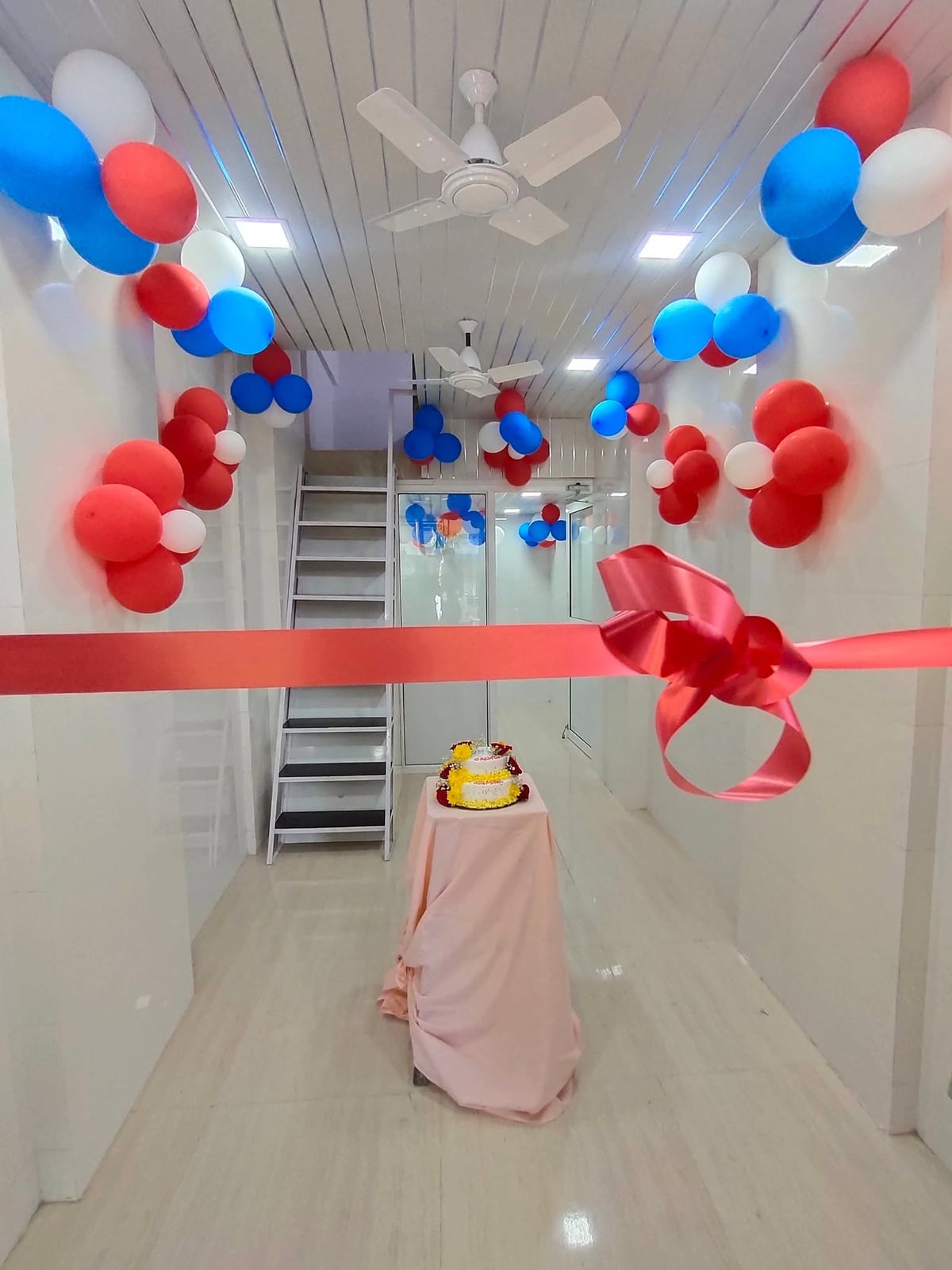 A ribbon waiting to be cut across a balloon-decorated hospital corridor, with a celebration cake on a draped table behind it.